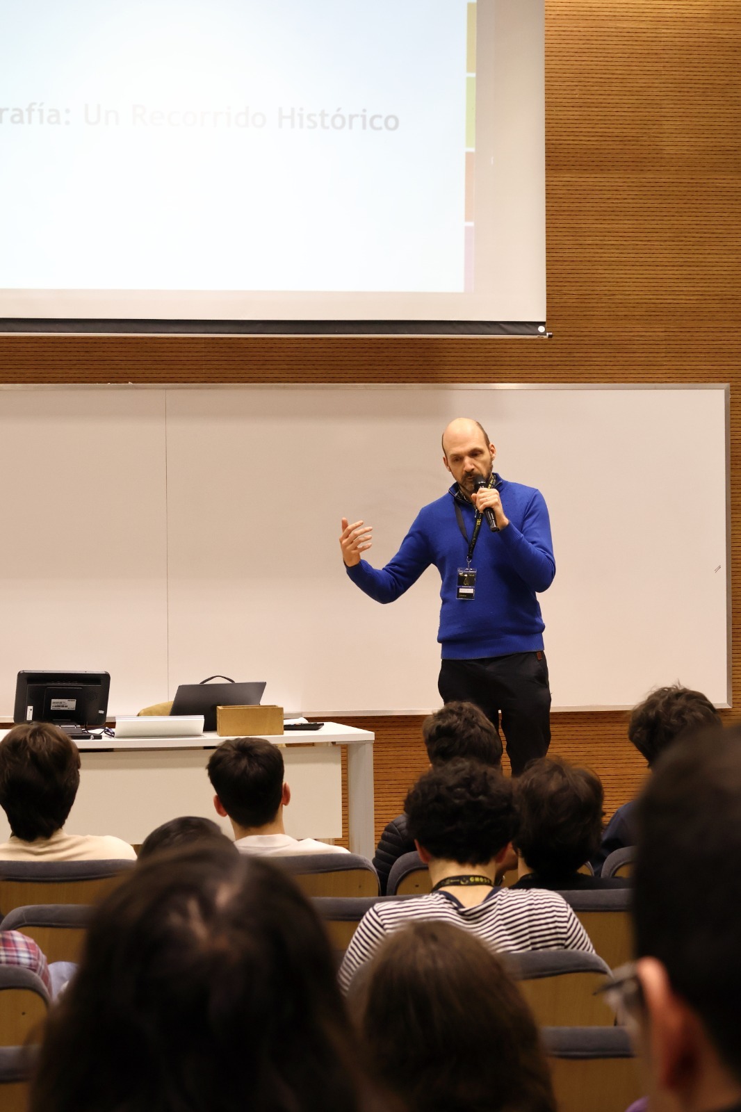 Pablo Deymonnaz holding a microphone at the front of the room, below a projected slide titled "Criptografía: Un Recorrido Histórico" (Cryptography: A Historical Journey)