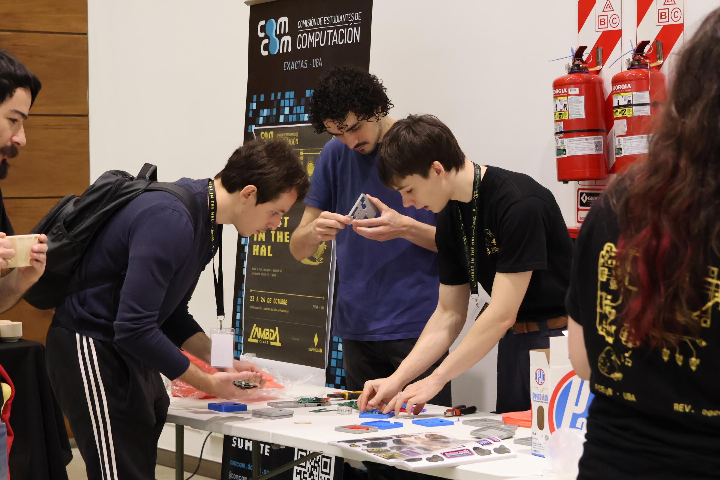 Ghost in the HAL participants assembling 3D-printed parts and boards on a table, in front of a banner of the Computer Science Students' Commission at Exactas UBA